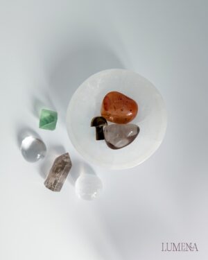 Image shows a Satin Spar (Selenite) bowl on a white background. In the bowl are a tumbled Smoky Quartz, a tumbled Carnelian crystal and a mini tiger's eye mushroom. Next to the bowl there's a Fluorite Octahedron, a tumbled clear quartz, a mini smoky quartz point and a satin spar (selenite) mini sphere.