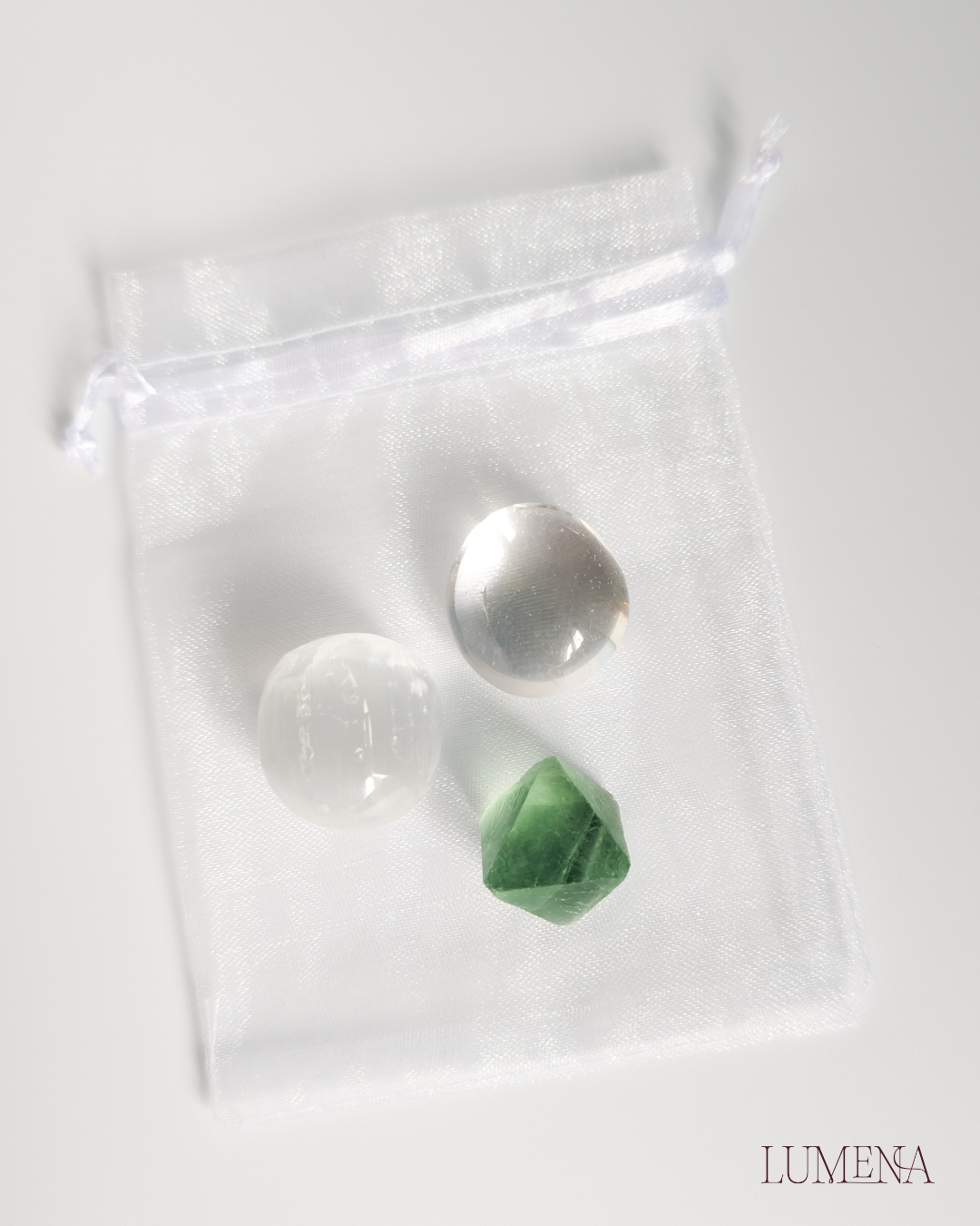 Image shows a small satin spar (selenite) crystal sphere, a clear quart tumble and a fluorite octahedron crystal collection, placed on a white table with a white organza bag.